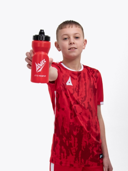 A kid is wearing a red football shirt with white panels and is drinking from a red water bottle with a self sealing nozzle