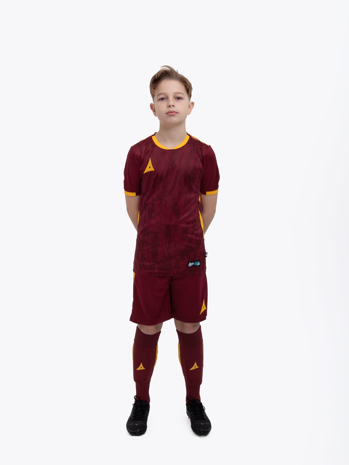 a kid standing up wearing a claret / maroon football shirt, matching claret shorts and claret football socks.